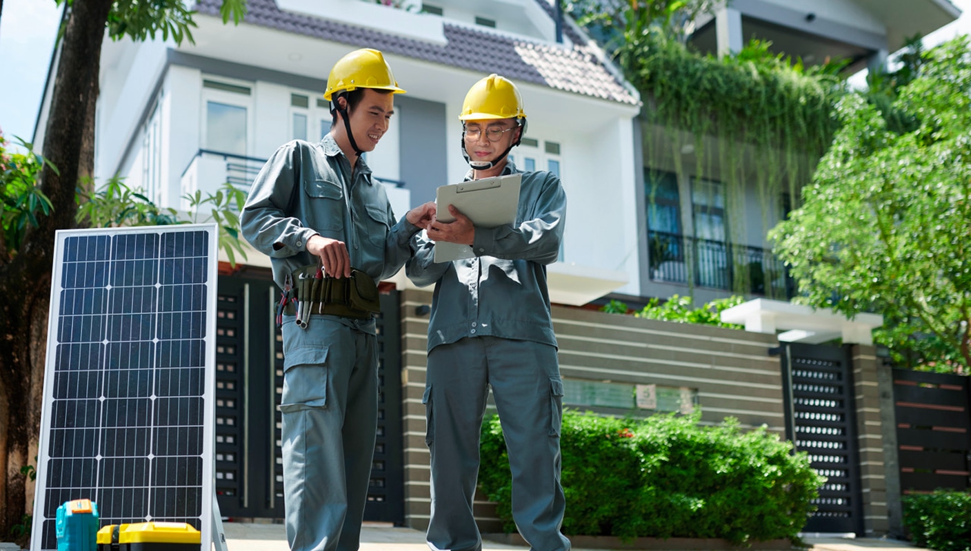 Two professionals in work attire reviewing documents on a tablet next to a solar panel, with a residential building in the background. The scene represents Solavita