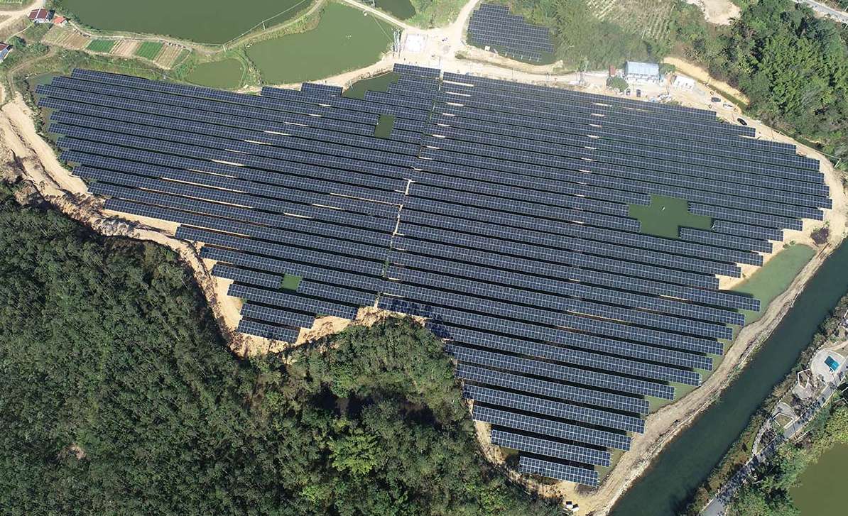 Aerial view of a large-scale ground-mounted solar power plant built on hilly terrain, showcasing rows of photovoltaic panels strategically placed around water bodies and uneven landscapes.