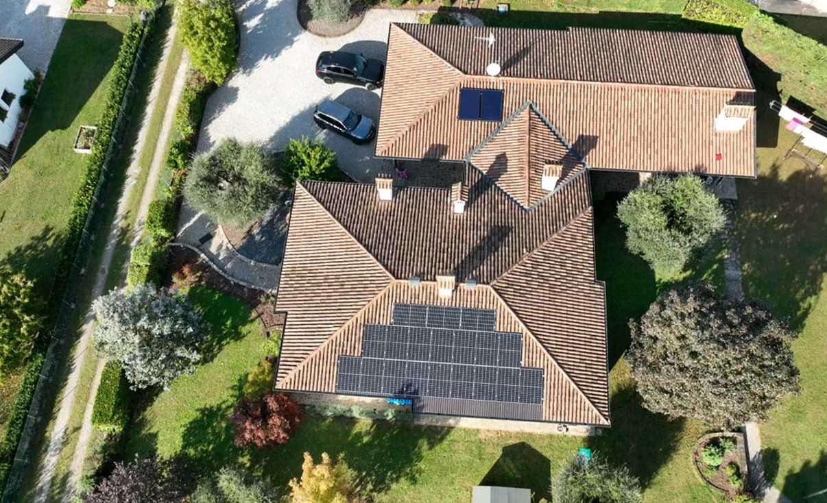 Aerial view of a modern European-style villa with neatly installed solar panels on a tiled sloped roof, surrounded by lush greenery and driveway access.