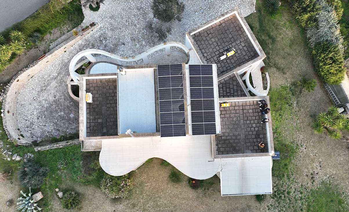 Aerial view of a modern villa with an architecturally artistic rooftop design and solar panels installed in two symmetrical rows, surrounded by stone pathways and landscaped greenery