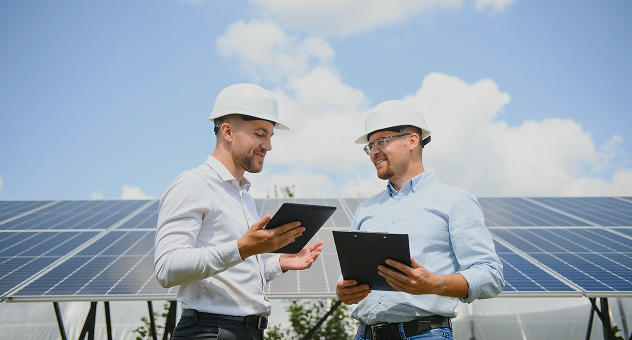 Two engineers discussing solar project details with tablet and clipboard at a photovoltaic site.