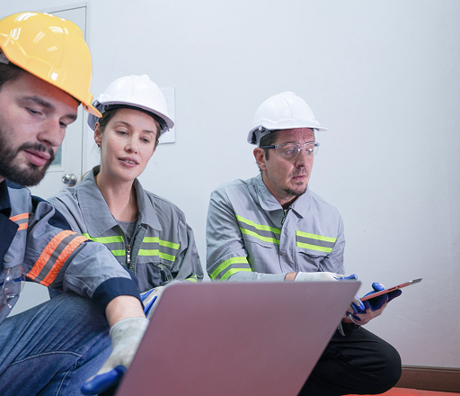 Three engineers wearing safety helmets and grey work uniforms are discussing something while reviewing data on a laptop and clipboard in a white indoor environment