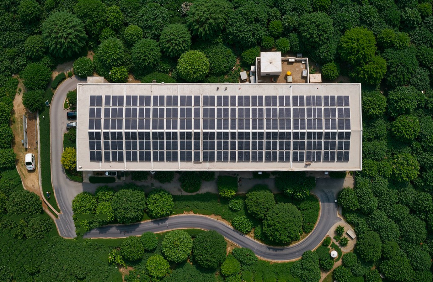 Aerial view of a building with solar panels installed on its roof, surrounded by greenery and a winding road
